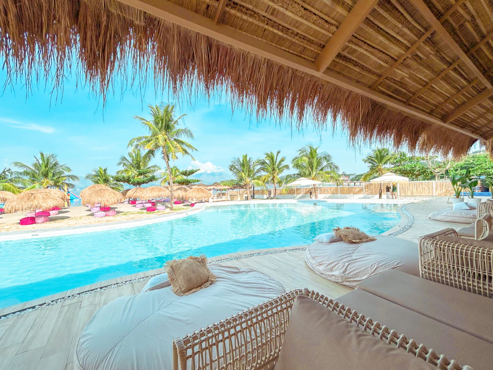 View of a pool with palm trees, lounge chairs, and umbrellas, seen from under a thatched roof structure with seating.