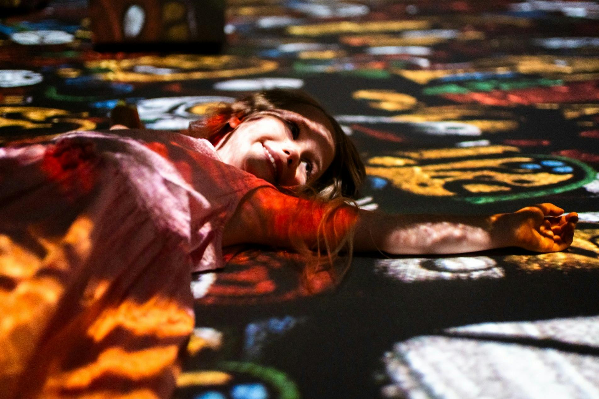 Smiling child lying on a colorful, patterned surface with light and shadow effects.