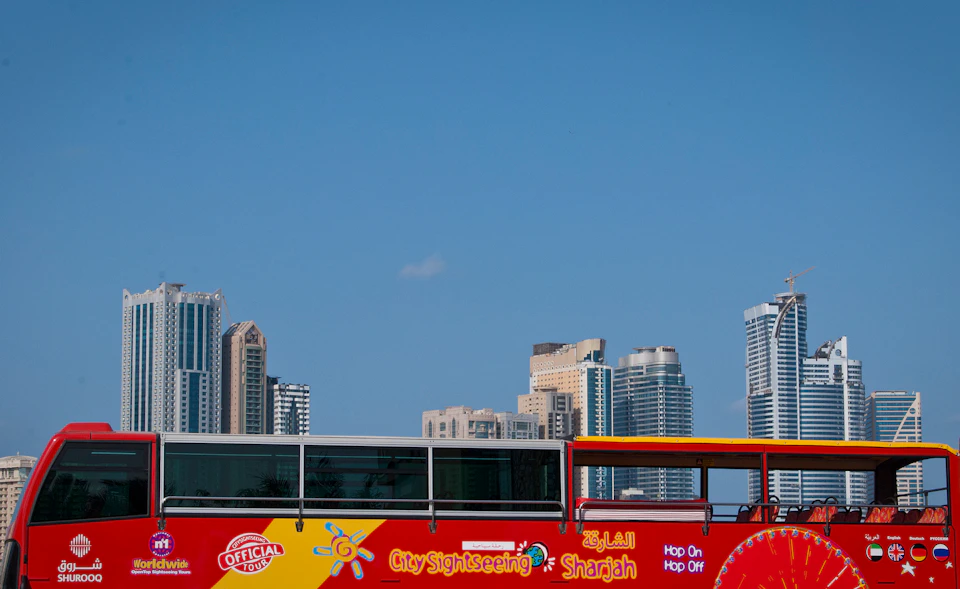 Red city sightseeing bus labeled "Sharjah" in front of tall modern buildings and a clear blue sky.
