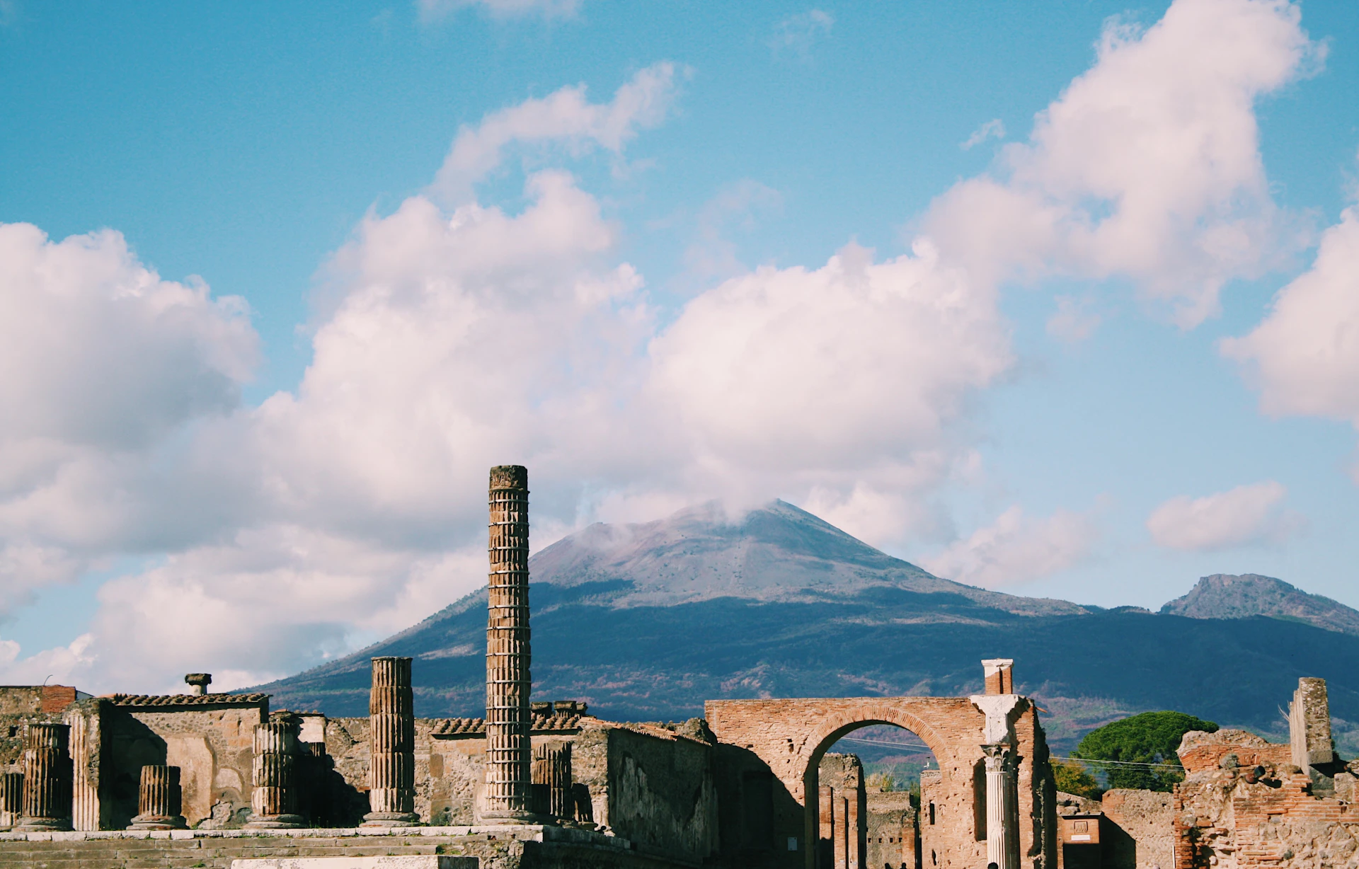 Ancient ruins with columns and arches under a blue sky, with a mountain in the background partially covered by clouds.