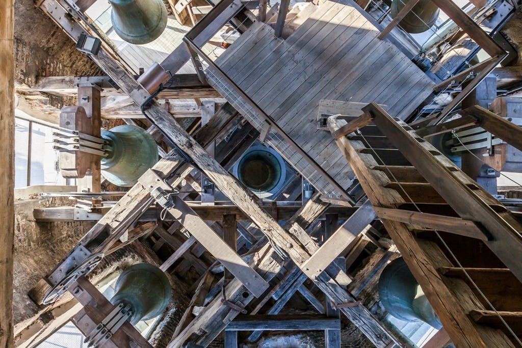 Interior of a bell tower showing multiple large green bells, wooden beams, and a wooden staircase.