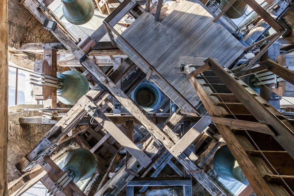 Interior of a bell tower showing multiple large green bells, wooden beams, and a wooden staircase.