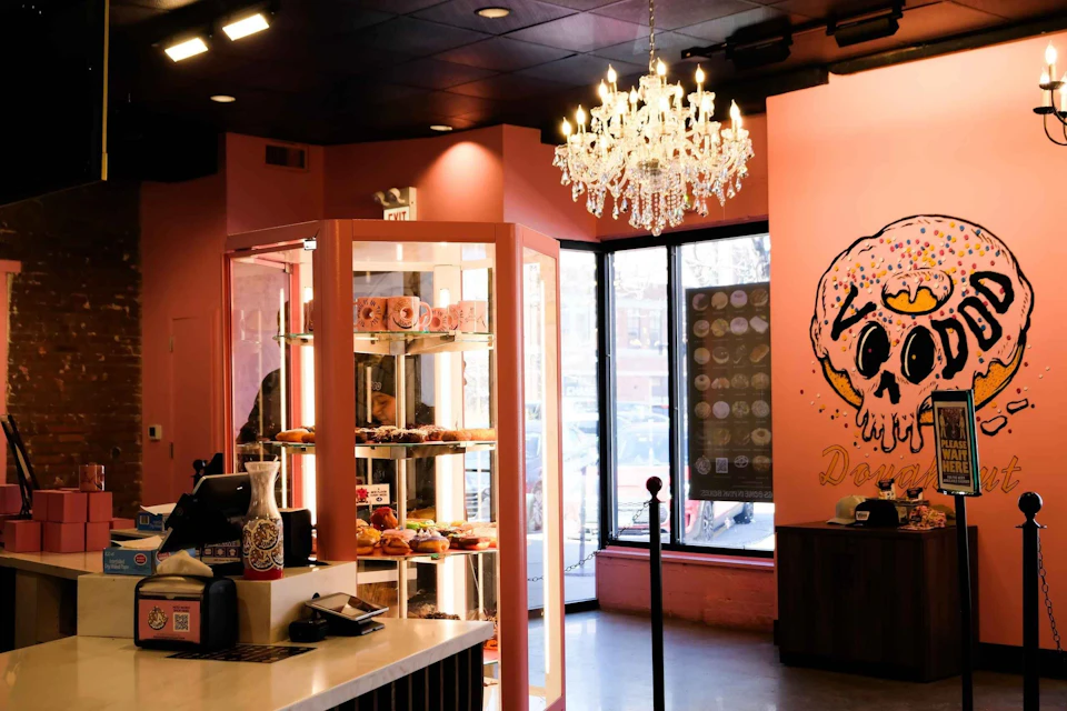 A cozy donut shop with pink walls, pastry display case, chandelier, and a large donut-themed mural near the window.