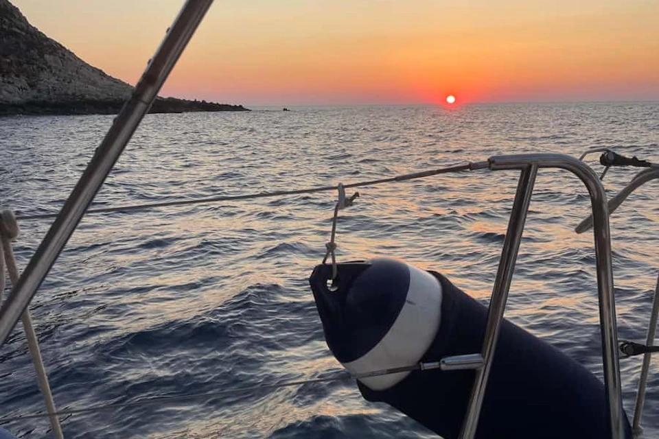 Sailboat railing and a blue and white fender with a sunset over a calm sea in the background.