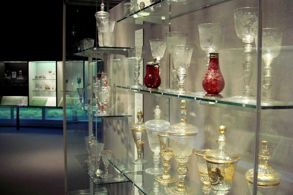 A display of ornate glassware and goblets, including red and gold decorative pieces, arranged on glass shelves in a museum.