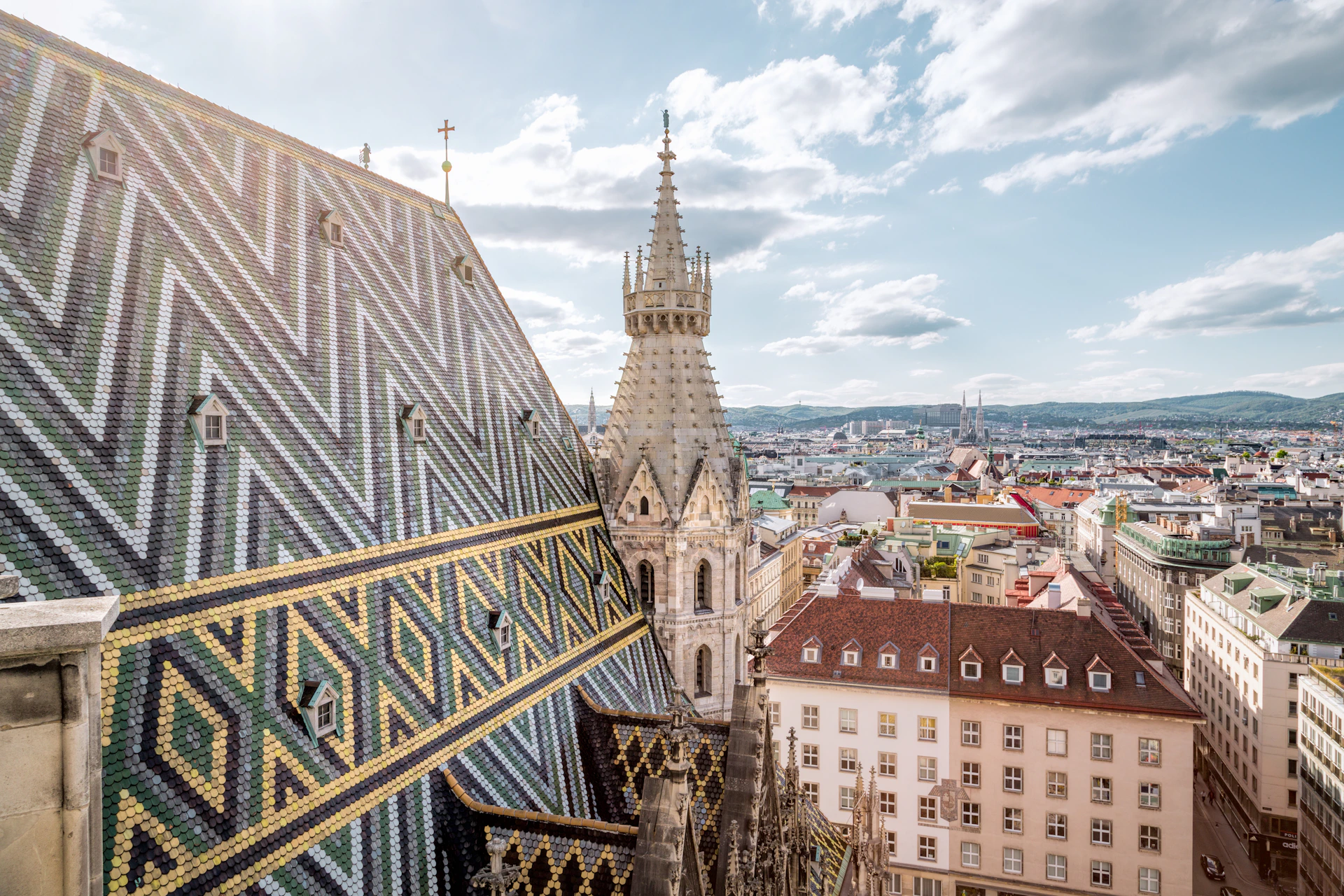 View of a cityscape with a decorative tiled roof, a tall, ornate spire, and surrounding historic buildings under a partly cloudy sky.