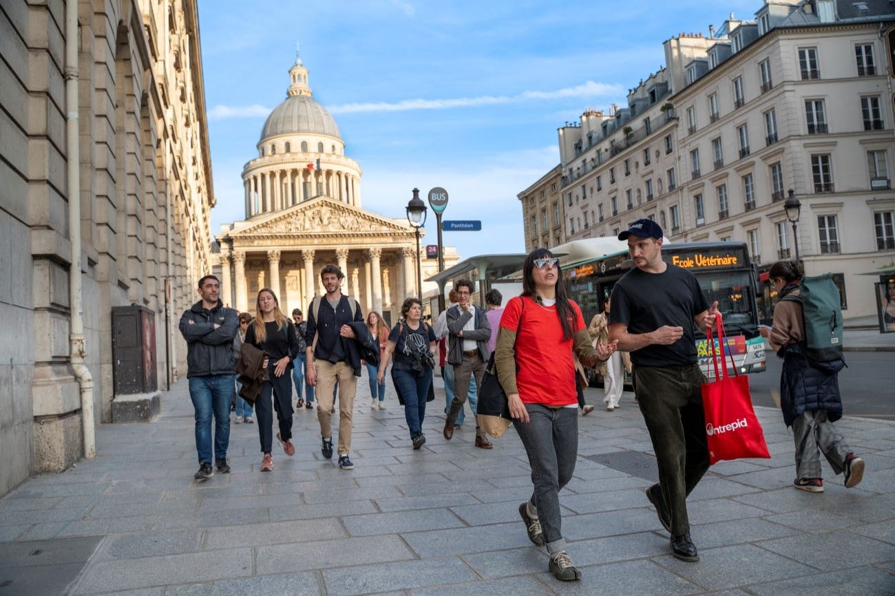 한 무리의 사람들이 푸른 하늘 아래 웅장한 돔형 건물인 판테온을 배경으로 도시 보도를 걷고 있습니다.