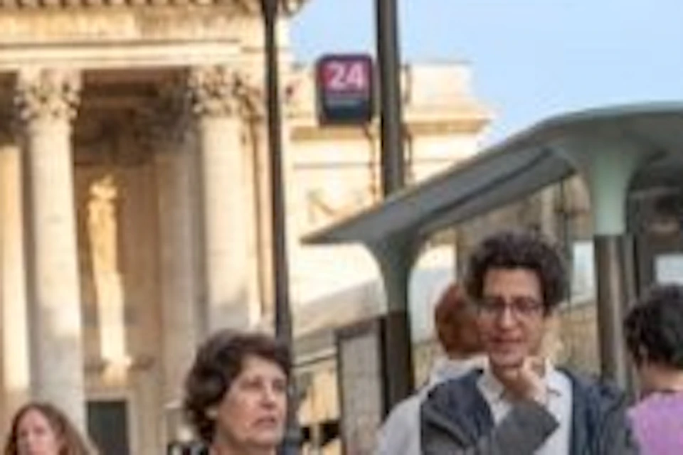 A group of people walk on a city sidewalk with the Pantheon, a grand domed building, in the background under a blue sky.