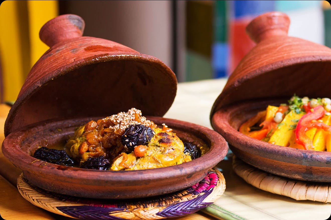 Two Moroccan tagines, one with a dish of chicken, prunes, and sesame seeds, the other partially visible with vegetables.