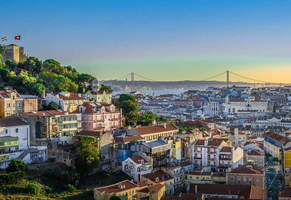 Colorful hillside buildings with a distant red suspension bridge over water, under a clear blue sky. Trees are scattered.