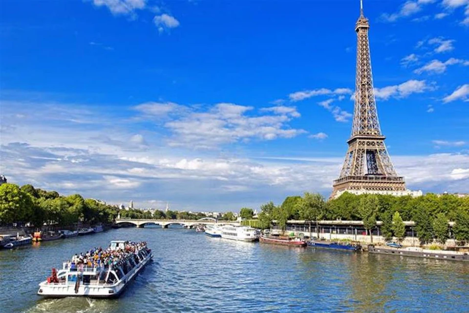 A boat with people floats on a river near the Eiffel Tower on a sunny day, with a bridge and greenery in the background.