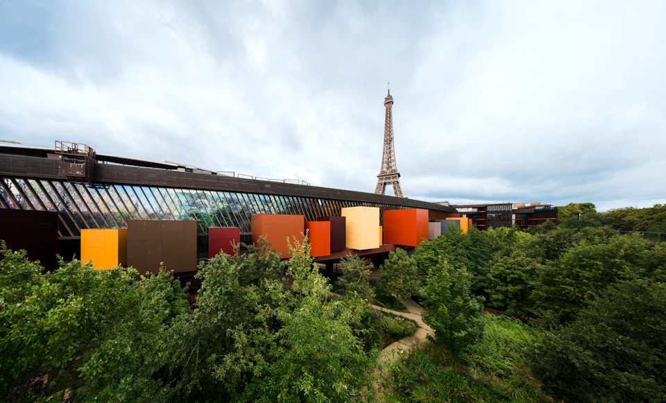 Colorful modern building surrounded by trees with the Eiffel Tower in the background under a cloudy sky.