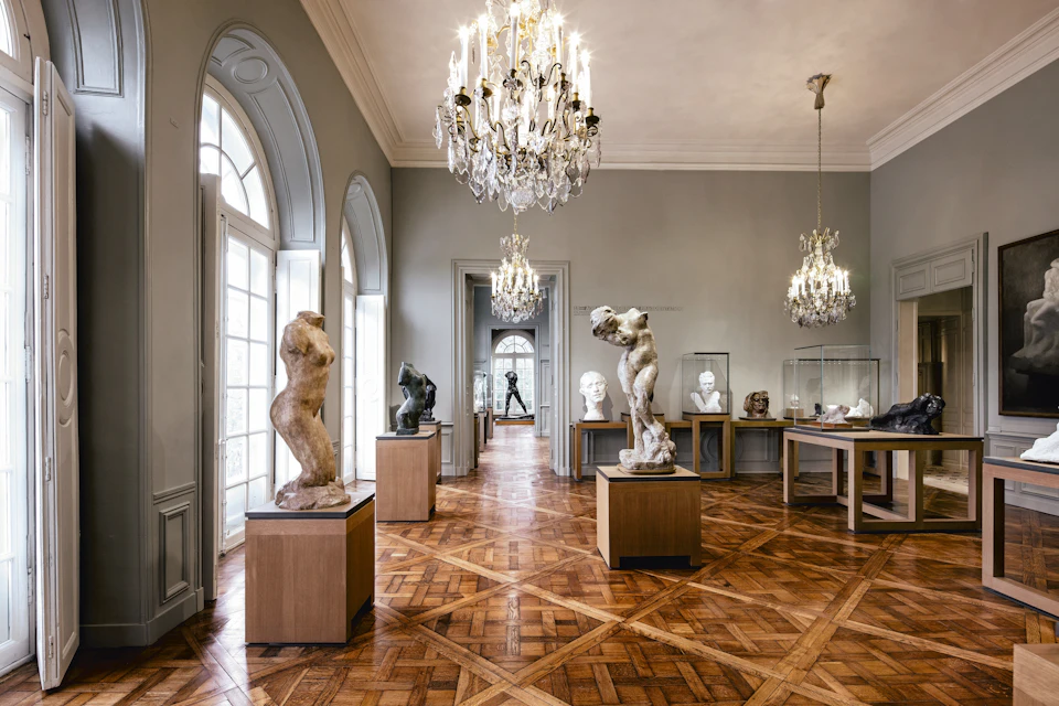 Gallery with wooden floors, chandeliers, and classical sculptures on pedestals. Busts and statues are displayed around the room.
