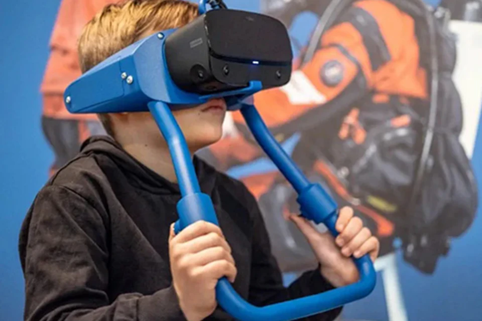 A person wearing a virtual reality headset with a blue frame and holding attached handles, against a blurred colorful background.