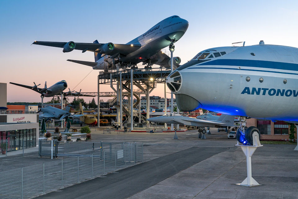 An outdoor aviation museum with various aircraft on display, including a large plane elevated on a platform at sunset.