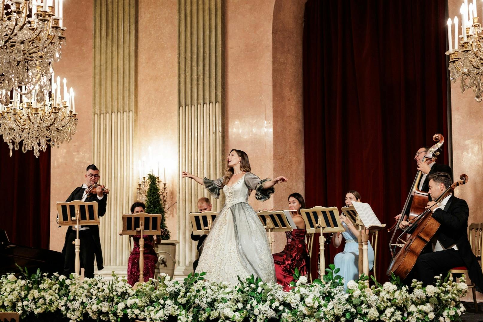 A woman in a period costume sings on stage with a small orchestra, surrounded by floral decorations in an elegant interior.