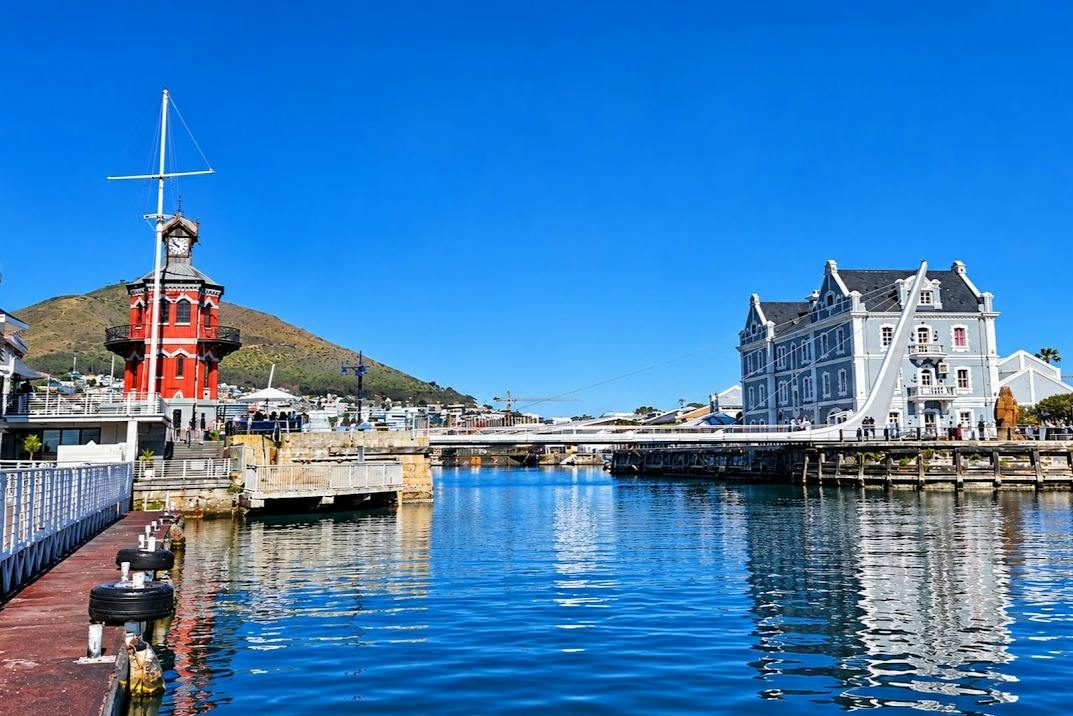 A waterfront scene with a red building, a gray building, a bridge, and a mountain in the background under a clear blue sky.
