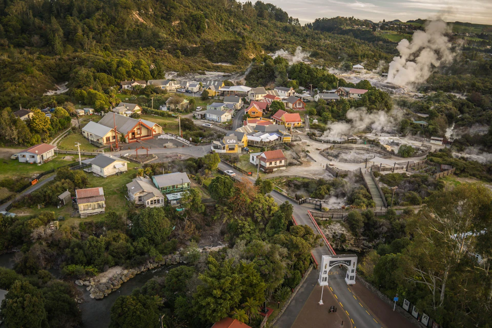 Aerial view of a village surrounded by greenery and hills, with steam rising from geothermal areas and buildings visible.
