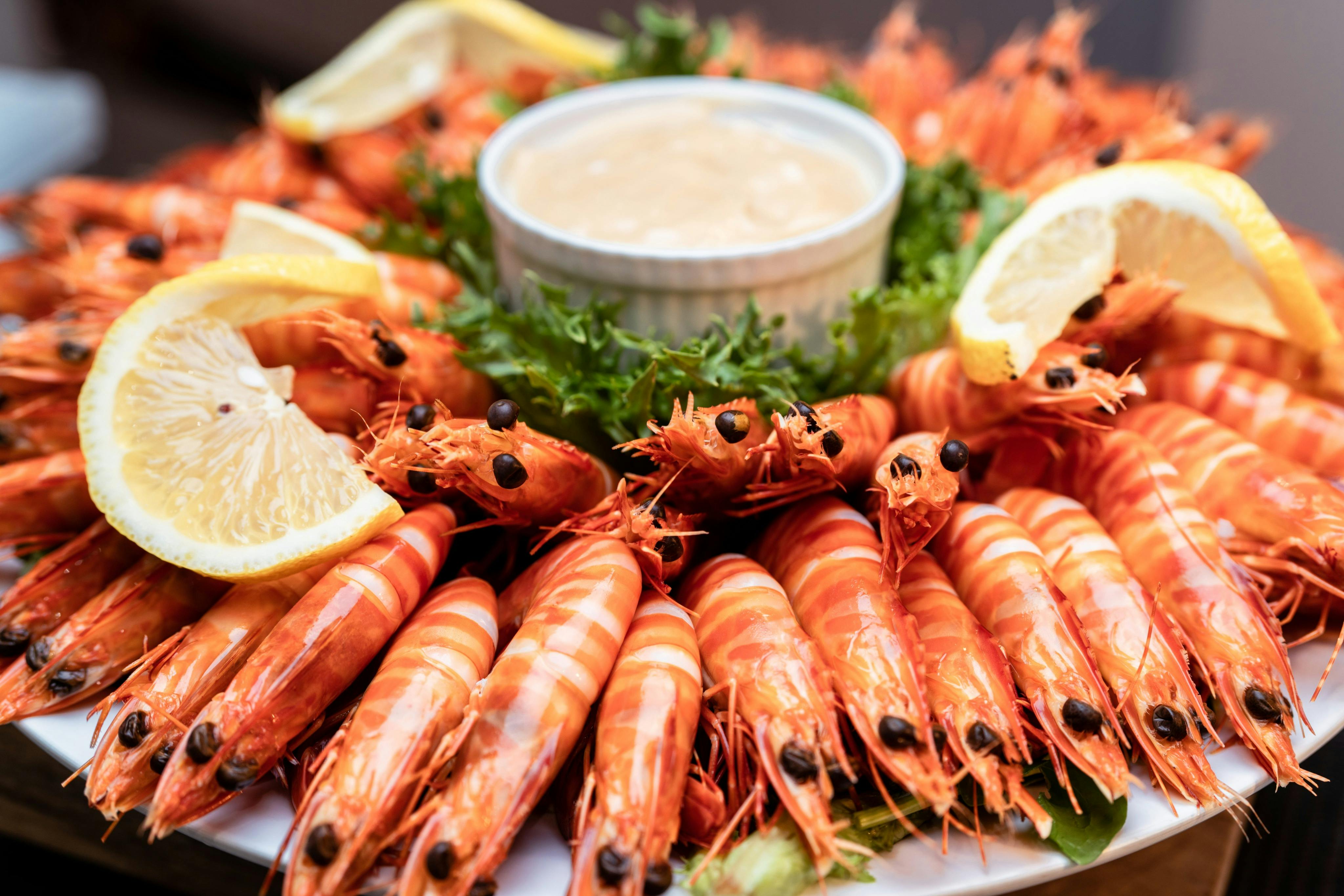 A platter of cooked shrimp arranged around a bowl of dipping sauce, garnished with lemon wedges and greens.