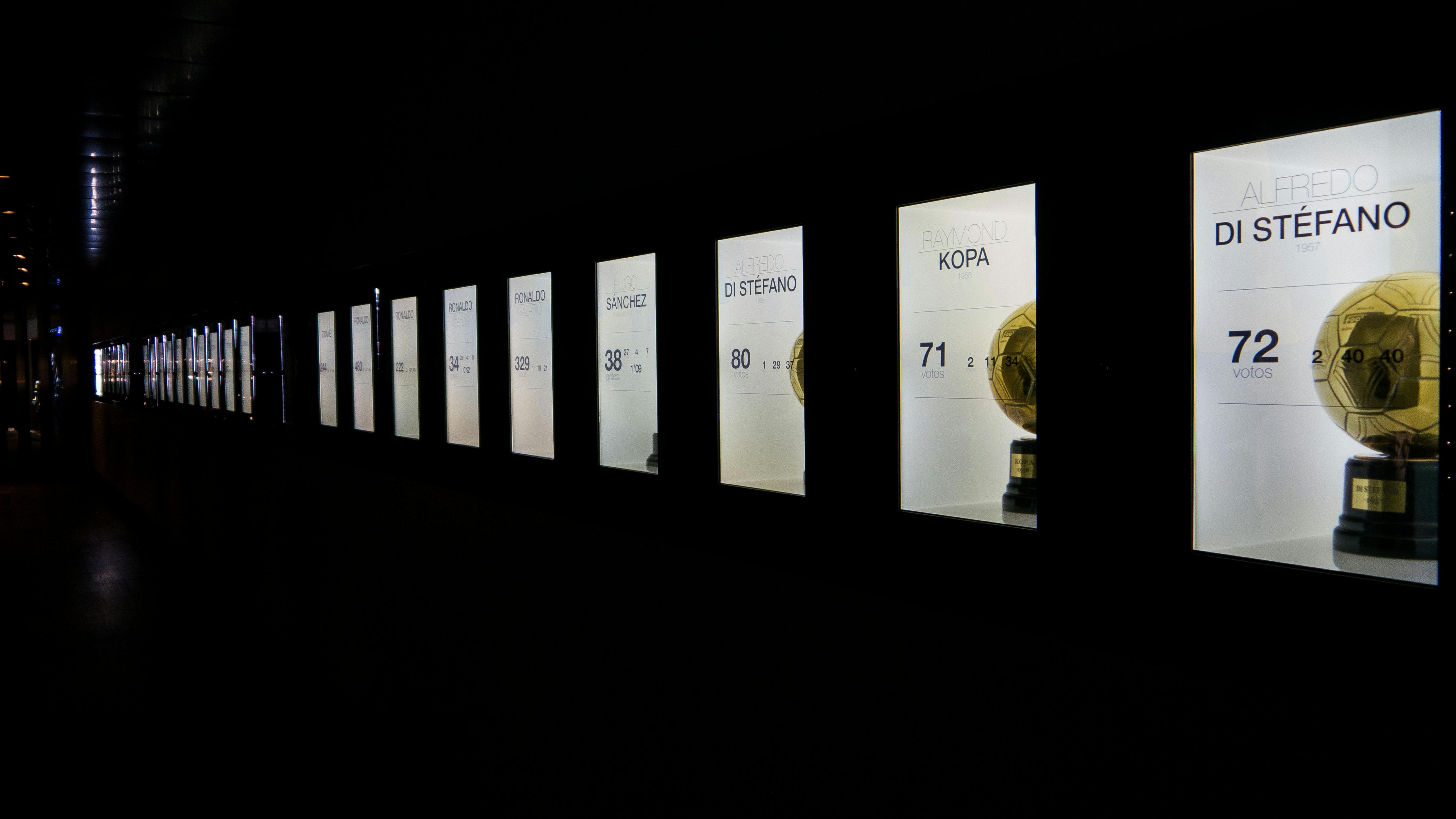 一排发光的展品展示了金色的足球奖杯，白色背景上有伴随的名字和数字。