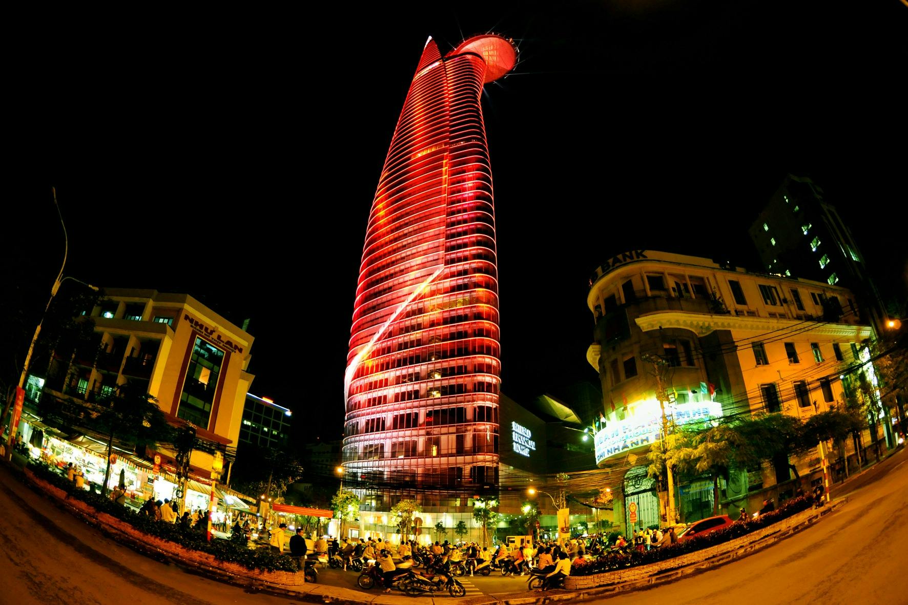 Futuristic skyscraper lit with red lights at night, surrounded by people, motorcycles, and other buildings in a busy urban setting.