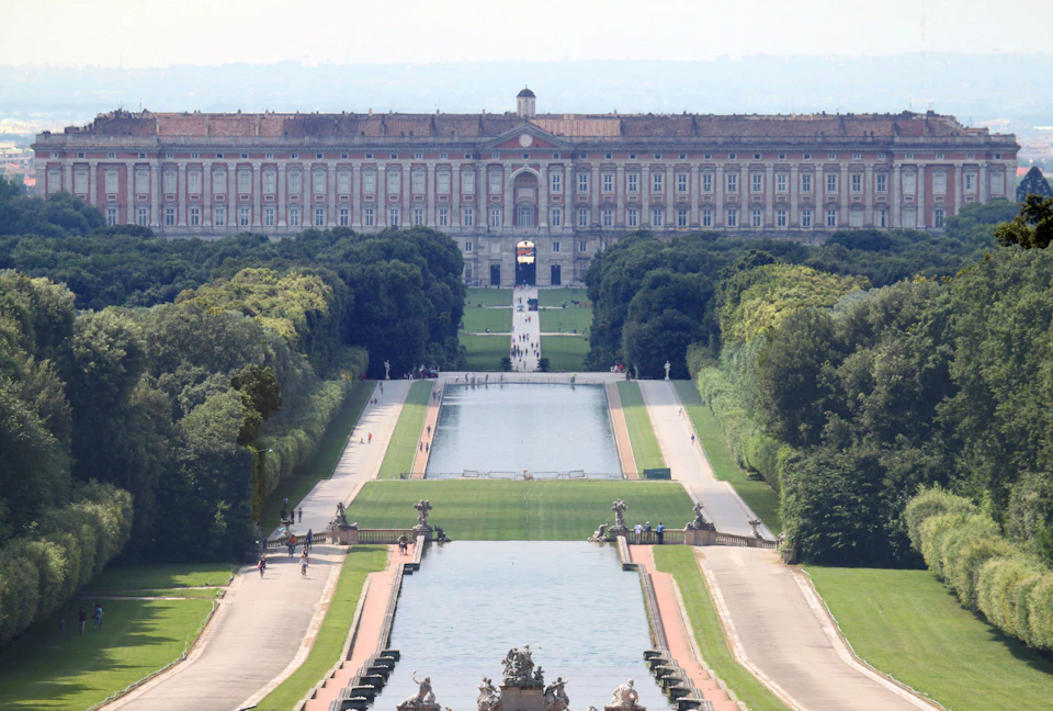 A grand palace with an expansive formal garden, featuring a long reflecting pool, statues, and tree-lined walkways.