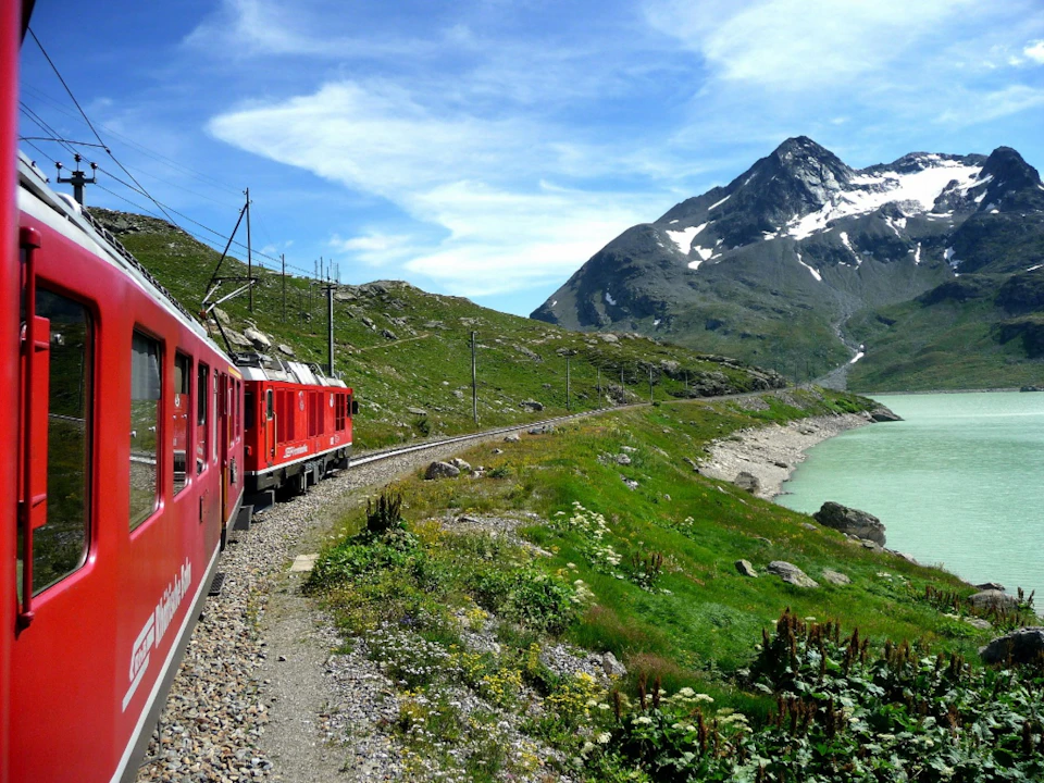 A red train travels along tracks next to a green lake, with mountains in the background under a blue sky with clouds.