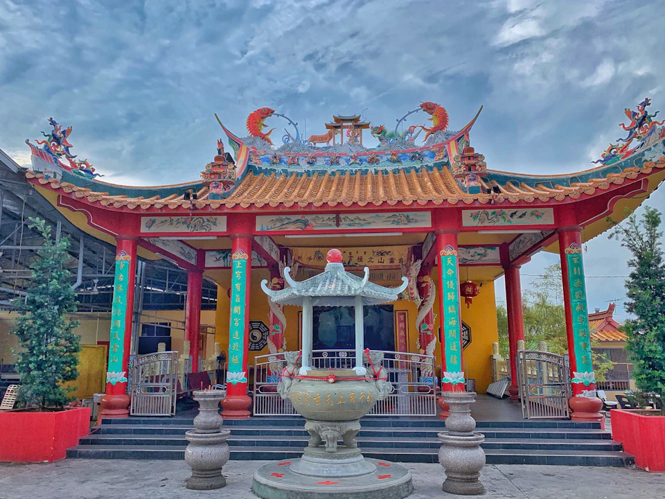 A traditional Chinese temple with ornate roof designs, red columns, and a large incense burner in front, under a partly cloudy sky.