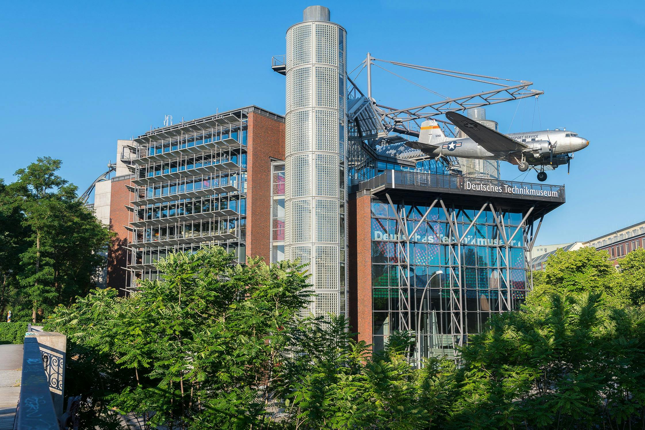 A modern building with a glass facade, a suspended airplane, and trees in the foreground on a sunny day.