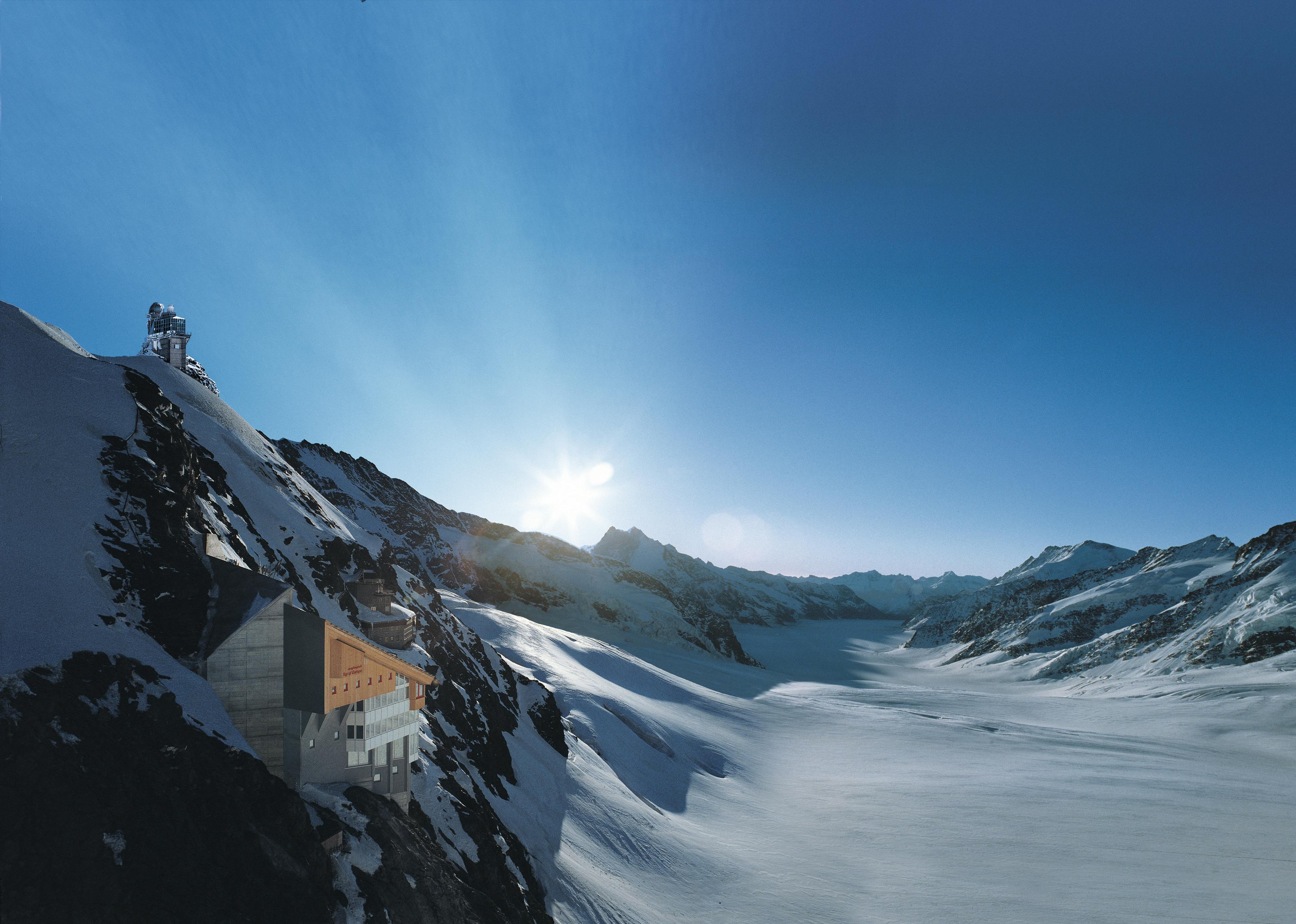 Zasněžená horská krajina s budovou posazenou na útesu a sluncem vycházejícím nad vzdálenými vrcholy.
