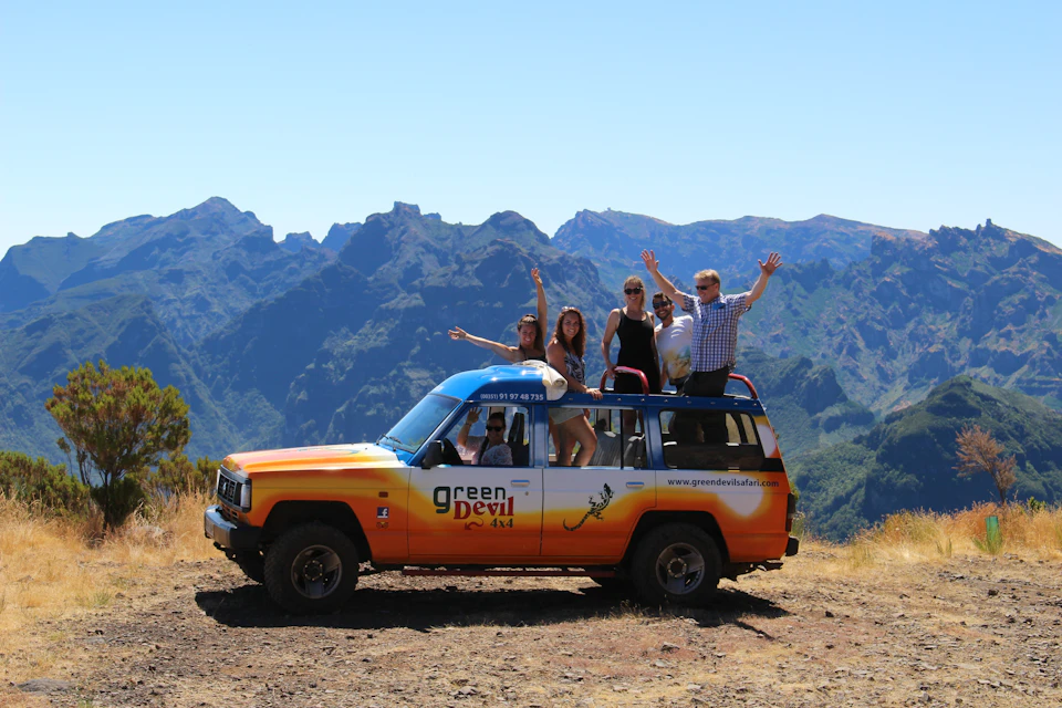 Five people standing and sitting on a colorful "Green Devil" tour jeep, with mountainous terrain in the background.