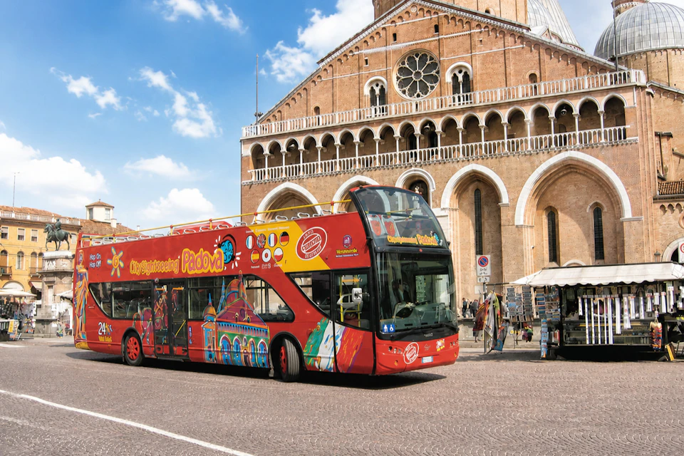 A red double-decker tour bus in front of a historic church with arched windows and a large rose window under a blue sky.