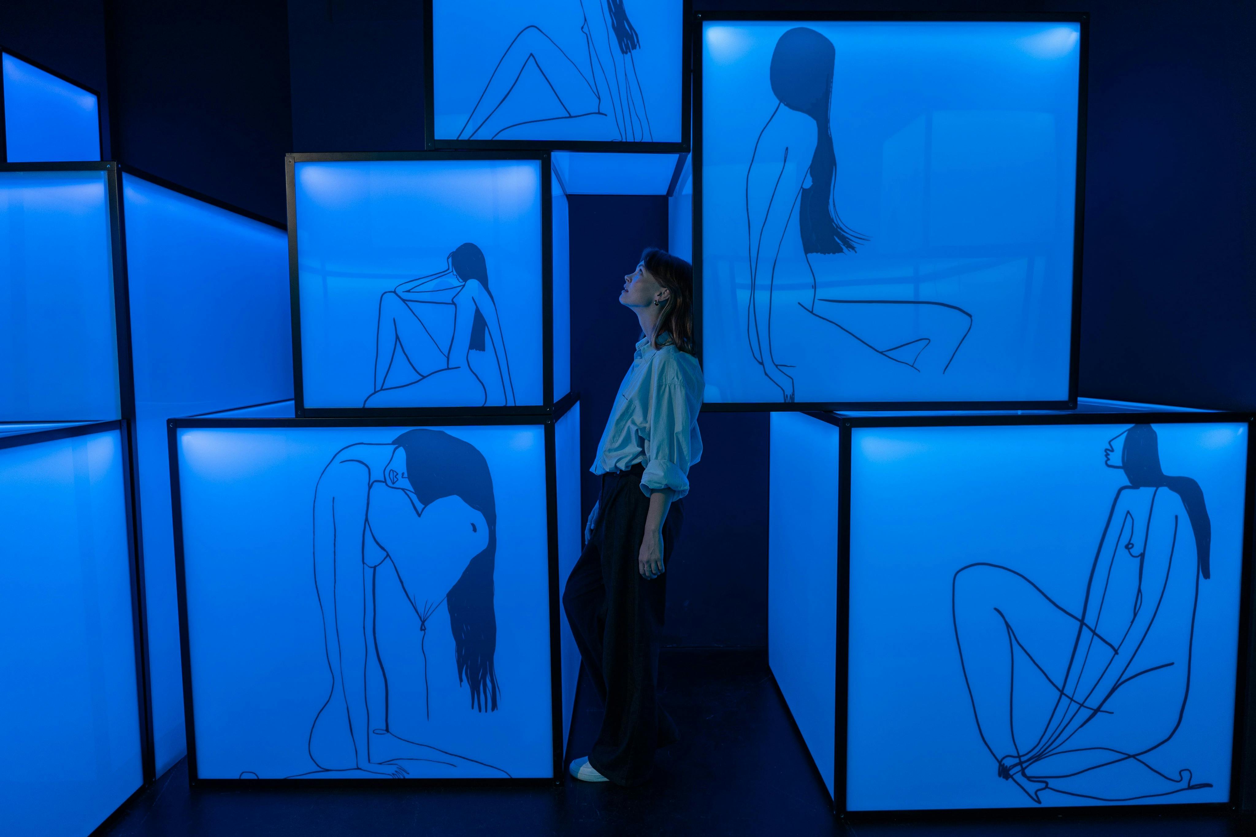 Obok ustawionych w stos podświetlanych niebieskich ekranów stoi osoba z minimalistycznymi rysunkami linii różnych siedzących postaci ludzkich.