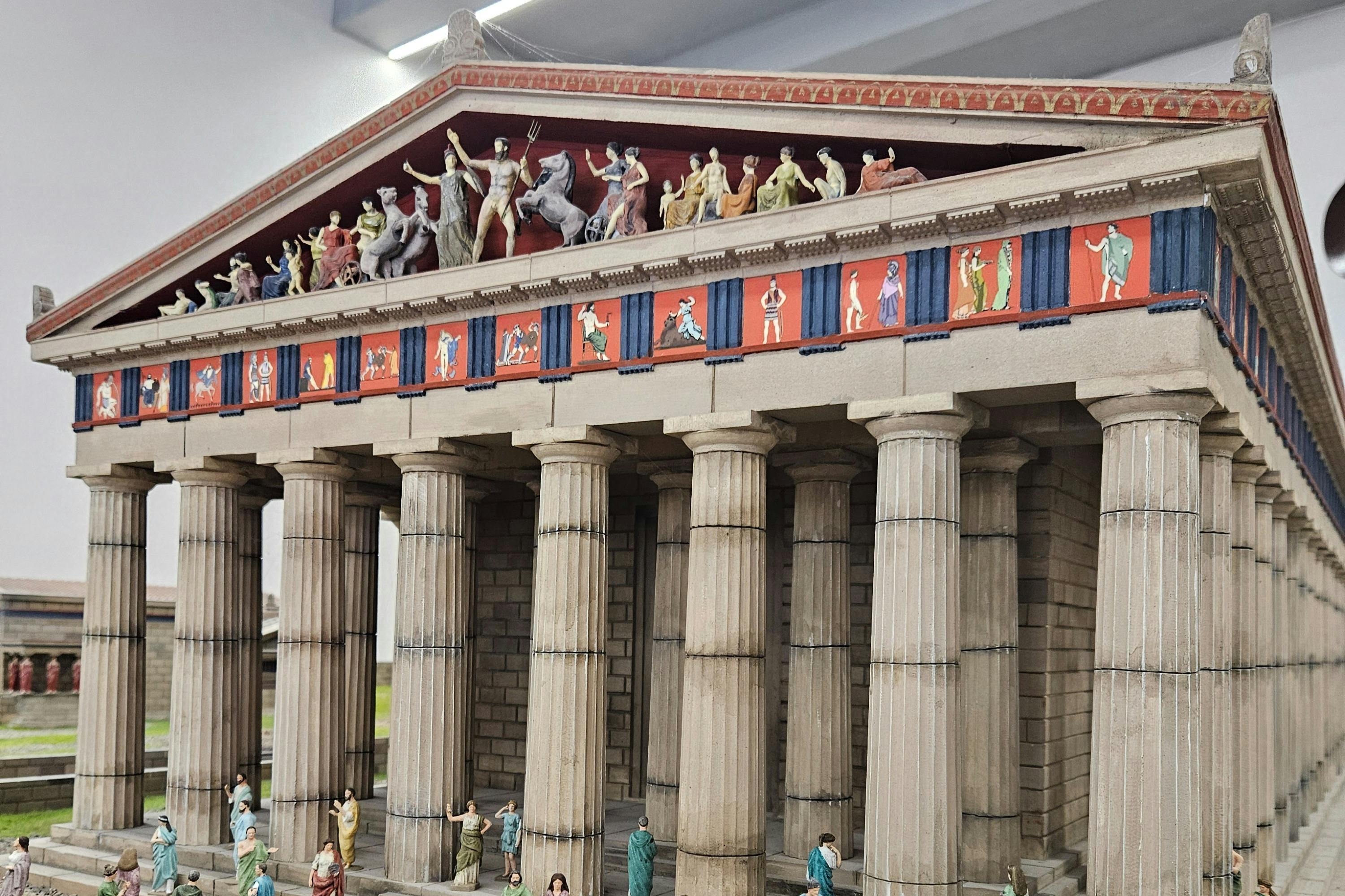 A detailed model of an ancient Greek temple featuring decorative sculptures and columns, with tiny human figures at the base.