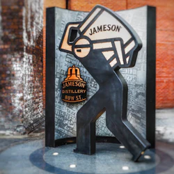 Skulptur av en stiliserad figur som håller en stor Jameson-flaska, med Jameson Distillery Bow St.-logotypen, mot en tegelvägg.