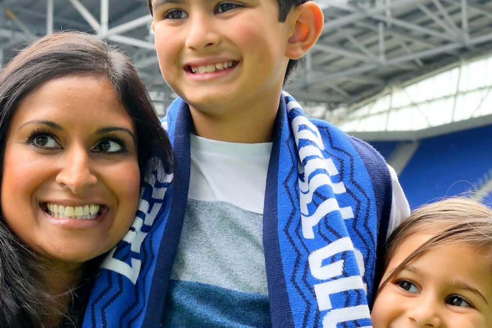 A woman and two children take a selfie together on the grass inside a large stadium, with a screen displaying "Tours."