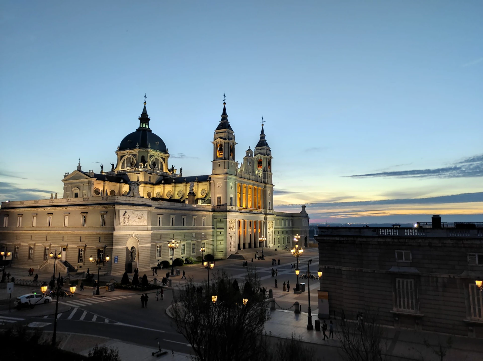 A large cathedral with two towers is illuminated at sunset. People walking and streetlights are visible in the foreground.