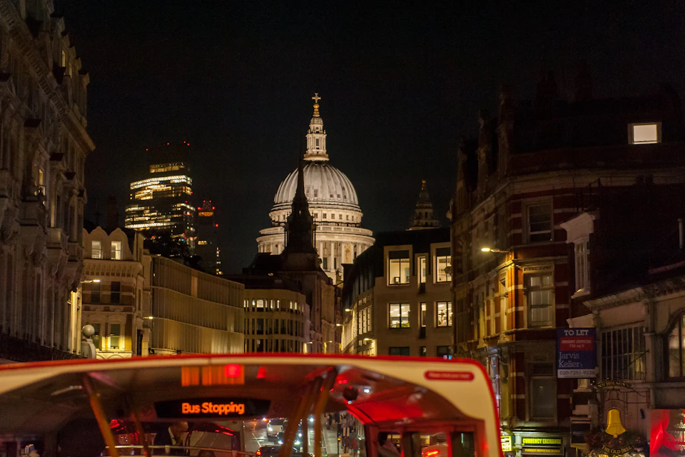 Nighttime cityscape featuring St. Paul's Cathedral dome illuminated, with a red double-decker bus in the foreground.