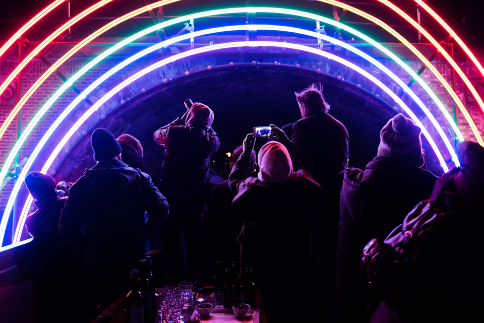 People in winter clothing taking photos under neon arches at night.