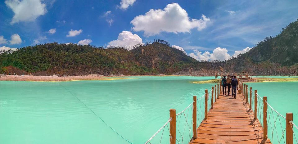 A turquoise lake surrounded by a forested hill under a blue sky with clouds, next to a wooden path with a rope railing.