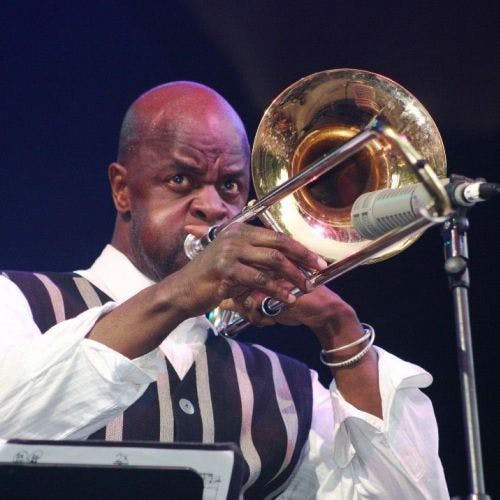 Muž intenzivně hraje na mosazný pozoun do mikrofonu, má na sobě pruhovanou vestu a bílou košili.