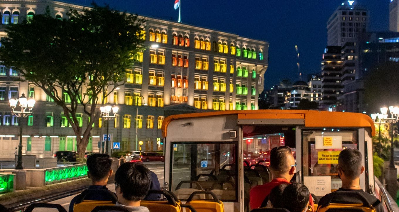 Lidé, kteří v noci jezdí autobusem s otevřenou střechou a míjejí jasně osvětlenou budovu s okny v zelené, žluté a oranžové barvě.