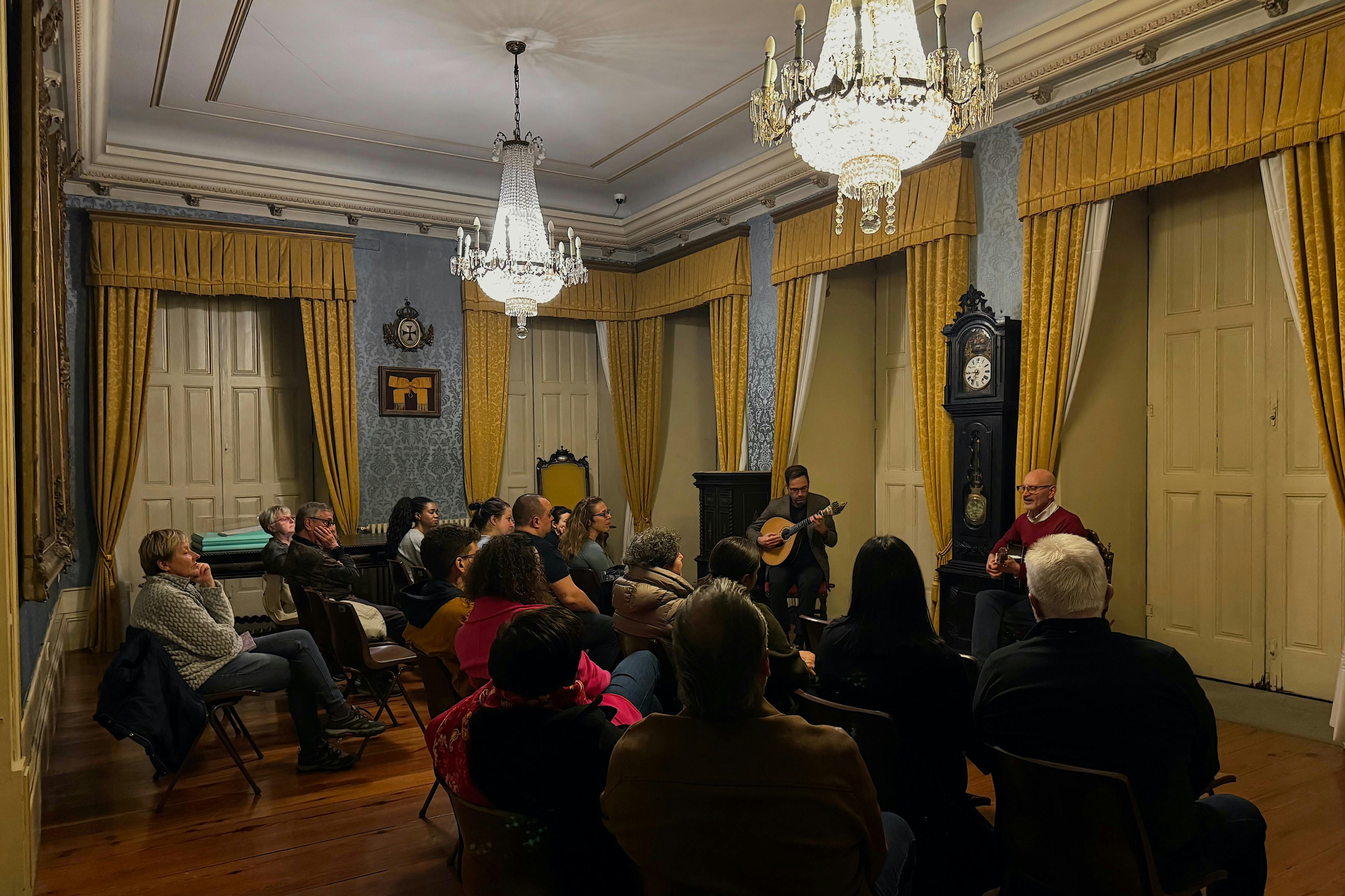 Skupina lidí sedících ve společenské místnosti s lustry a poslouchající dva hudebníky hrající na kytary.