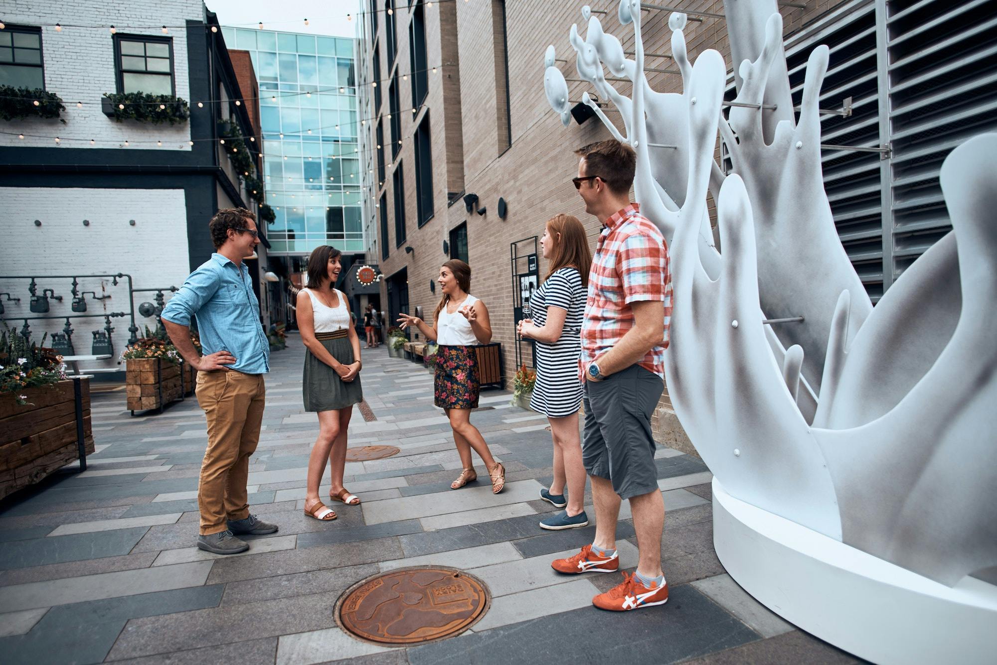 Un groupe de cinq personnes discutant dans un espace urbain moderne, en plein air, avec des œuvres d'art décoratives, des plantes et des bâtiments autour d'eux.