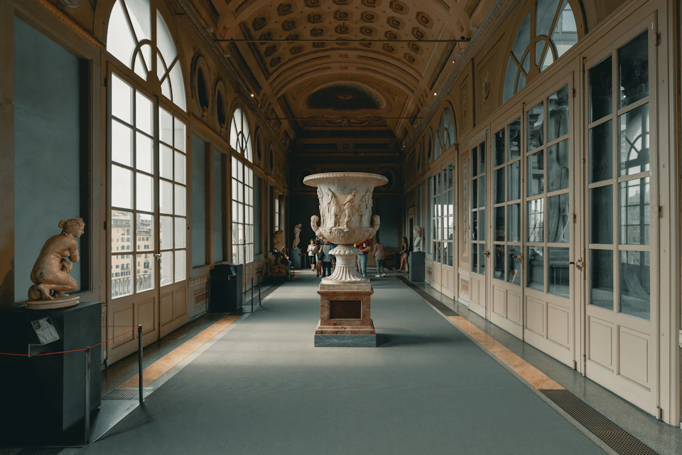 Ornate gallery with tall windows, statues, and a large decorative vase in the center. People are seen in the background.