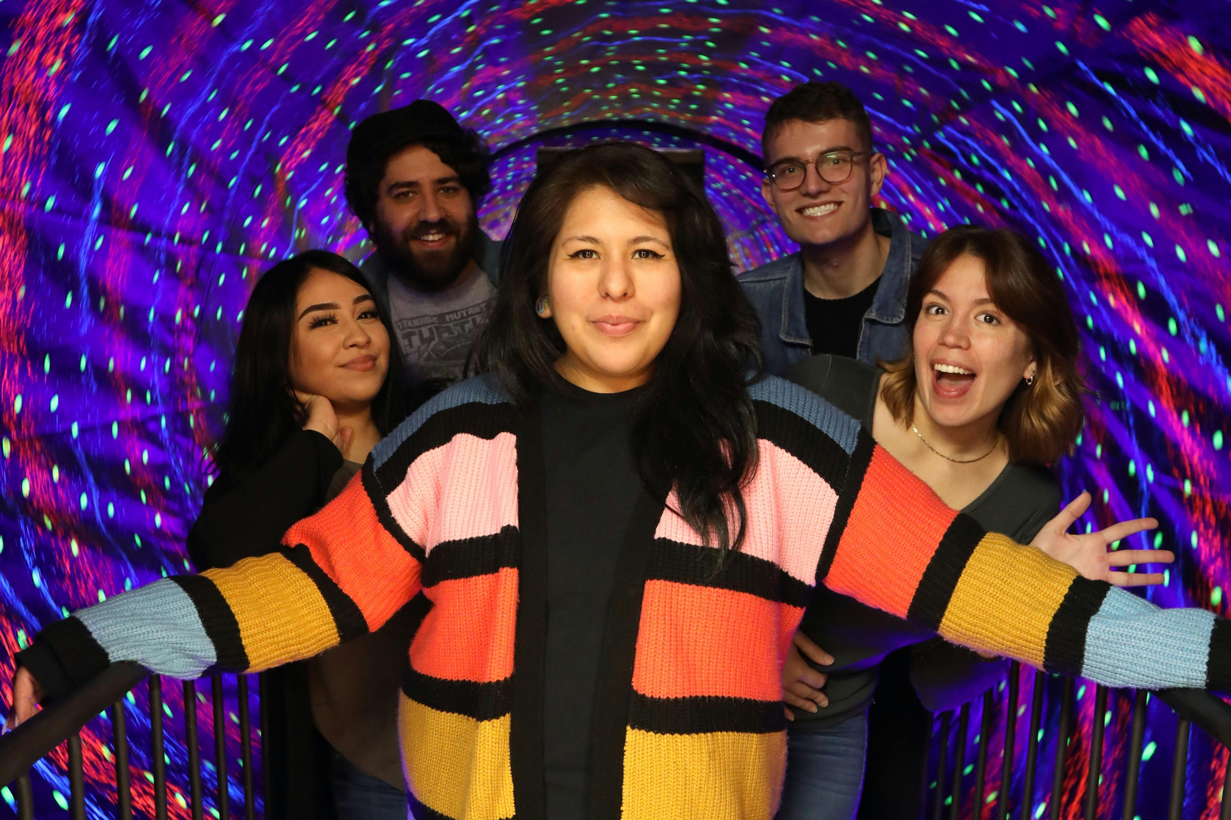A group of five people smiling and posing in a tunnel with colorful, swirling lights in the background.