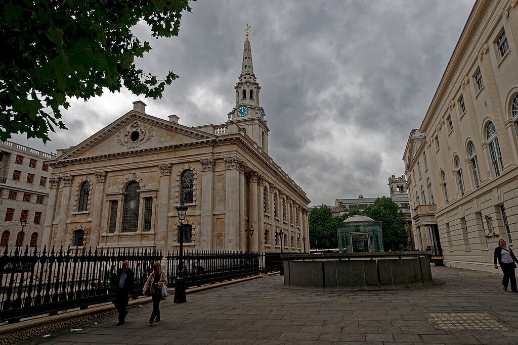 一座历史悠久的教堂，多云的天空下有一座钟楼，周围环绕着古老的建筑物。 人们在铺好的街道上行走。