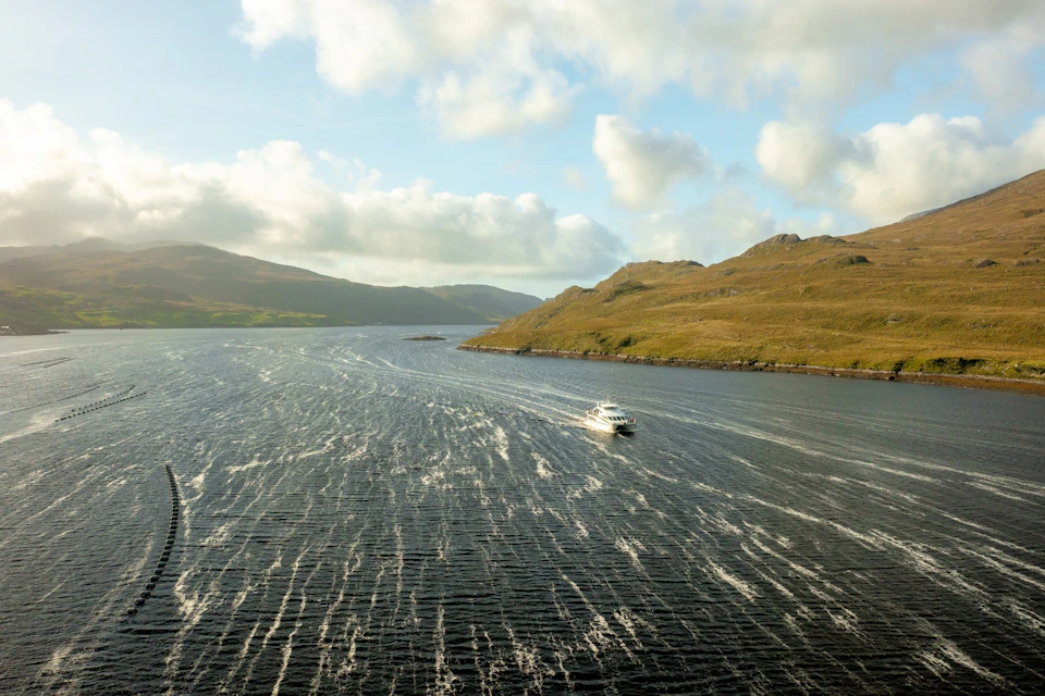 A ferry navigates a scenic, wide river surrounded by rolling hills under a partly cloudy sky.