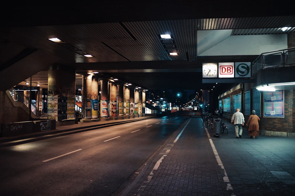 A quiet street at night illuminated by streetlights, with a single person walking past a building displaying a clock and "DB" sign.