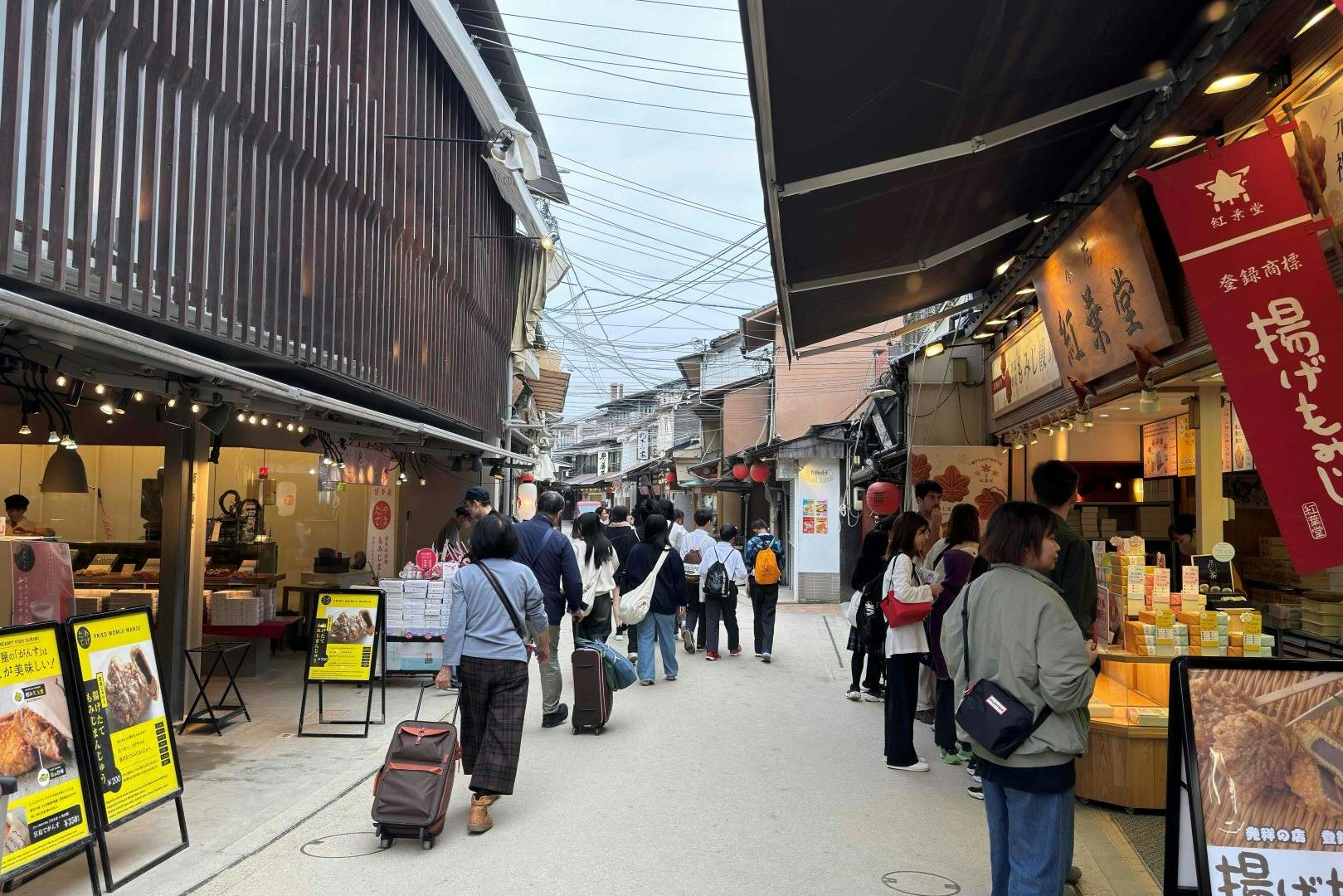 A bustling street market with people walking and shopping at stalls with food and goods under awnings and signage.